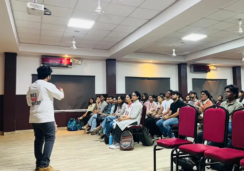 A speaker in a Build Theory shirt addresses students seated in a hall at an AI Readiness Session